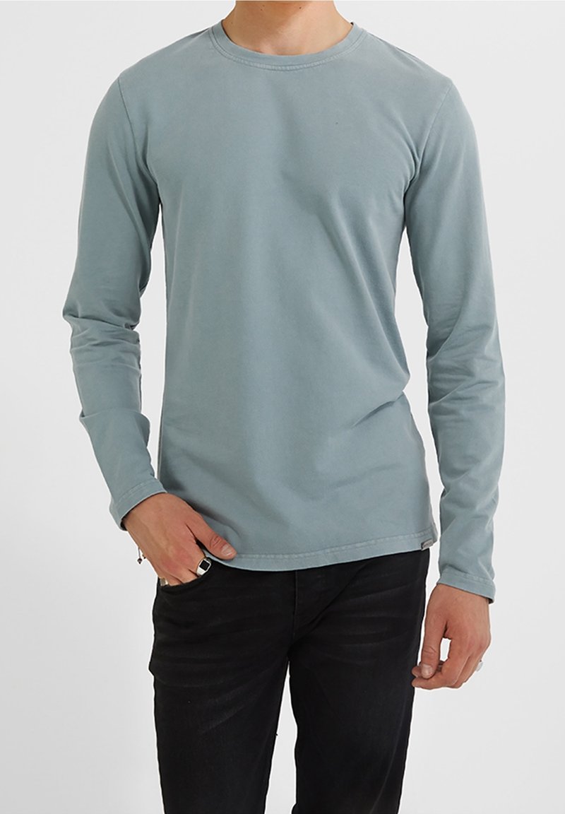 Homme portant une chemise ajustée bleu clair à manches longues et un pantalon noir avec une bague noire à la main droite.