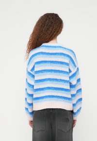 Πλεκτό πουλόβερ με οριζόντιες ρίγες σε μπλε και ροζ, μαλακή υφή, στρογγυλό ντεκολτέ και μακριά μανίκια· φορεμένο πάνω από γκρι παντελόνια με τσέπες.