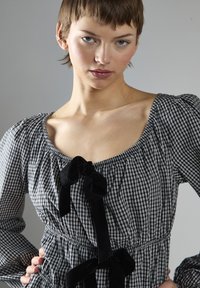 Joven con cabello corto vistiendo una blusa a cuadros en blanco y negro con lazos de terciopelo negro, con las manos en las caderas, posando frente a un fondo gris liso.