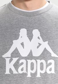Grå sweatshirt med et fremtrædende hvidt motiv af to siddende silhuetter med krydsede ben samt "Kappa"-logoet nedenunder.