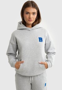 Šedá mikina vyrobená z měkkého materiálu, má přední kapsu a vyšívané modré písmeno 'B' na hrudi. Jednoduchý design s kapucí.