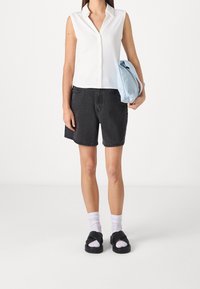 Femme portant un haut sans manches blanc à boutons, un short en denim noir, des chaussettes blanches avec des sandales noires, tenant une veste légère bleu clair repliée.