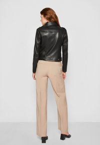 Giacca in pelle nera con design aderente, collare a contrasto e fibbie laterali; abbinata a pantaloni alti beige chiaro e scarpe con tacco a blocco.
