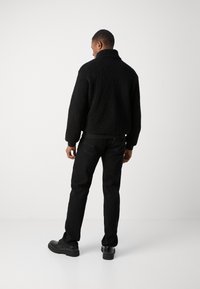 Giacca di pile nera con colletto alto, abbinata a jeans scuri e scarpe nere. Materiale strutturato visibile sulla superficie della giacca.
