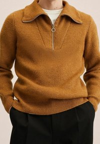 Strickpullover in einem warmen Senfton mit einem Reißverschluss an einem Stehkragen, gerippter Textur und einer taillierten Silhouette. Kombiniert mit dunkler Hose.