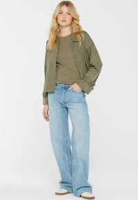 Olivgrüner Bomberjacke über einem olivgrünen Hemd, kombiniert mit hellblauen weitgeschnittenen Jeans und beigen Sneakers, vor einem weißen Hintergrund stehend.