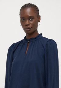 Maglietta blu navy a maniche lunghe realizzata in tessuto leggero. Presenta un colletto arricciato e una piccola apertura a forma di chiave sul davanti.