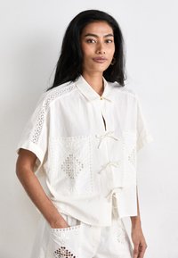 Camicia bianca a maniche corte con motivi ricamati su spalle e tasche, con chiusura a bottoni e dettagli con cordoncino.