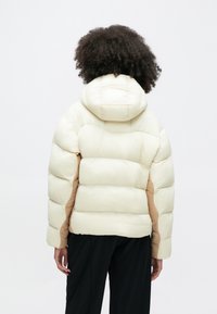 Osoba o kręconych włosach w kremowej puchowej kurtce z kapturem i beżowymi bocznymi panelami, stojąca tyłem na jednolitym białym tle.