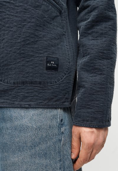 Μπλε ναυτικό τζάκετ με υφή και καμπυλωτή λαιμόκοψη, με ετικέτα κεντημένη "PS Paul Smith" στο κάτω αριστερό μέρος. Συνδυάζεται με ανοιχτόχρωμο τζιν παντελόνι.