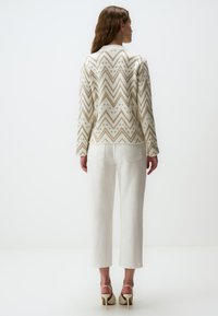 Brede gebreide cardigan in crème met een gouden zigzagpatroon. Heeft lange mouwen en een ronde halslijn. Gecombineerd met witte rechte jeans.