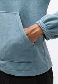 Χέρι ατόμου μέσα στην μπροστινή τσέπη ενός ανοιχτό μπλε φλις πουλόβερ με ελαστικά μανίκια, εμφανές πάνω σε λευκό φόντο.