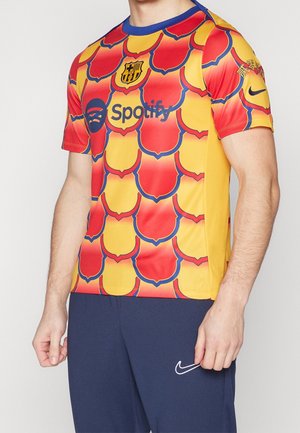 Man met een rood-gele schubbenpatroon sporttrui met clublogo, Spotify als sponsor en zwarte Nike-broek.