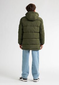 Grüne Daunenjacke mit Kapuze und gestepptem Design. Kombiniert mit hellblauen Jeans und schwarzen Turnschuhen, von hinten betrachtet.