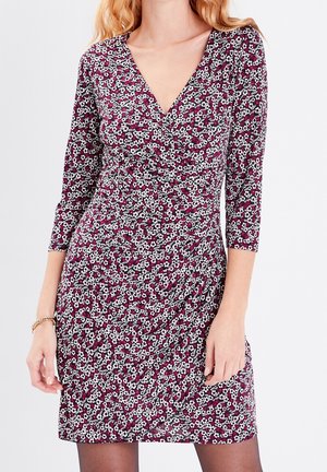 Femme portant une robe ajustée à imprimé floral avec manches trois-quarts et décolleté en V, debout devant un fond clair uni.