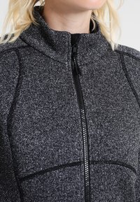Woman wearing a black and grey textured zip-up jacket with high collar, shown from neck to mid-torso against a plain background.