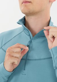 Vaaleansininen, pitkähihaisella neljännesvetoketjulla varustettu pulloveri, jossa on sileä pinta, litteät saumat ja osittain avattu vetoketjukaulus.