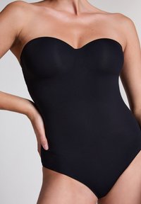 Vrouw die een zwarte strapless badpak met een sweetheart-halslijn draagt en poseert met één hand op haar heup.
