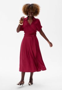 Bordeaux wikkeljurk met korte mouwen, textuurstof, zelfstrik taille en vloeiende rok. Modellen dragen gepatenteerde hakken en accessoires.