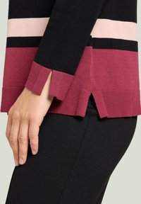 Strickpullover mit vertikalen Streifen in Schwarz, Pink und Burgunderrot. Seitliche Schlitze am Saum und eine Bündchenverarbeitung. Weiche Textur und figurbetonte Form.