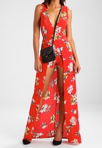 Tuta floreale rossa con scollo a V profondo e spacchi laterali, realizzata in tessuto leggero. Abbinata a una piccola borsa nera e tacchi neri.