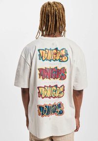 Světle béžové oversized tričko s výrazným, vícebarevným textem ve stylu graffiti na zadní straně, který opakovaně hlásá "DINGERS DANGEROUS".