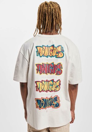 Light beige oversized t-shirt featuring bold, multicolored graffiti-style text on the back, stating "DINGERS DANGEROUS" in repeated layers.