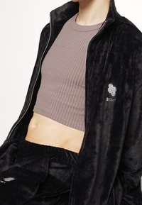 Zwarte velvet jas met rits en geborduurd logo, daaronder een grijze geribbelde cropped top met textuur; zacht, pluche stof; casual pasvorm.