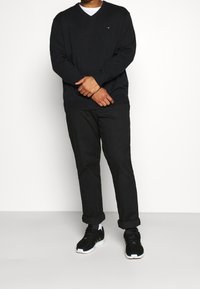 Pull à col en V bleu marine, pantalon noir et baskets noires. Le pull présente un petit logo, le pantalon est retroussé aux chevilles et les chaussures ont des accents blancs.