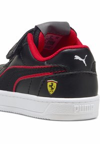 Zapatilla de cuero negra con acentos rojos, que cuenta con una correa de Velcro, suela blanca y un logo amarillo de Ferrari en el lateral. Logo de Puma en el talón.