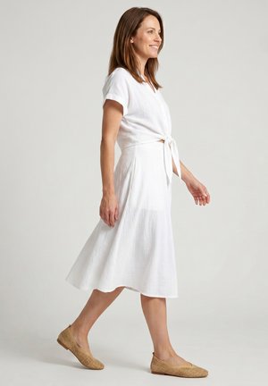 Vrouw in een witte korte mouw jurk met een voorsluiting, lopend en lachend, met lichtbruine geweven platte schoenen, tegen een effen lichte achtergrond.