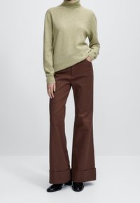 Hellgrüner Rollkragenpullover mit langen Ärmeln, kombiniert mit braunen, weit geschnittenen Hosen mit aufgekrempelten Bündchen und glatter Textur.