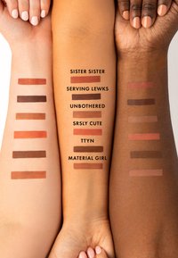 Quattro braccia che mostrano otto campioni di diverse tonalità, tra cui toni terrosi, etichettati con nomi. Texture liscia su pelle chiara e scura.