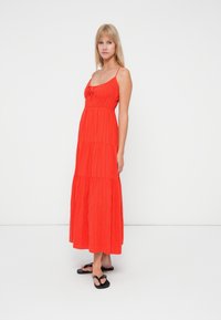Rode maxi-jurk met spaghettibandjes, gestructureerde stof, gelaagd ontwerp en een decoratieve strikdetail op de borst. Draagt met zwarte sandalen.