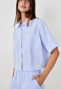 Camicetta corta a maniche corte in cotone a righe blu e bianche con due tasche sul petto e chiusura con bottoni davanti.