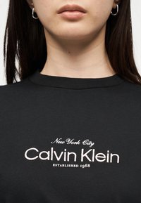 Κοντινό πλάνο ατόμου που φοράει μαύρο πουκάμισο με κεντημένο κείμενο στο στήθος: "New York City Calvin Klein Established 1968".