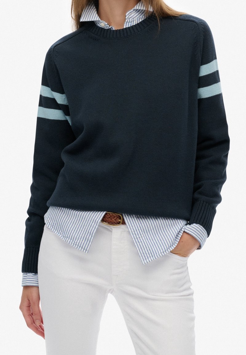 Personne portant un pull bleu marine avec deux rayures bleu clair sur chaque manche, sur une chemise rayée bleue et blanche boutonnée, associée à un pantalon blanc.