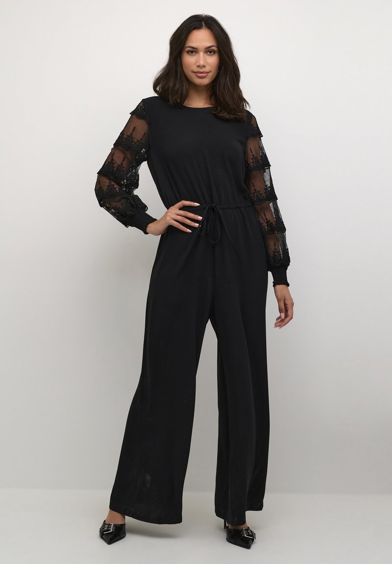 Schwarzer Jumpsuit mit durchsichtigen, langen Spitzenärmeln. Hat einen runden Halsausschnitt, eine Kordelzug-Taillierung und ein weit geschnittenes Design. Glatte Textur.