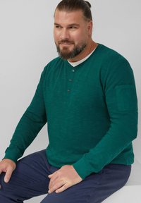 Ciemnozielona koszula z długim rękawem z fakturowanej tkaniny, z okrągłym dekoltem, listwą guzikową oraz kieszonką na lewym rękawie.