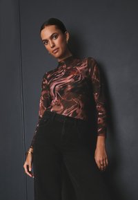 Kobieta ubrana w długorękawowy, transparentny brązowo-czarny wzorzysty top oraz czarne spodnie z wysokim stanem, stojąca na tle ciemnej ściany.