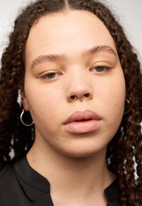 Gros plan d'une personne avec des cheveux bouclés brun moyen portant un t-shirt noir, des boucles d'oreilles en sautoir et plusieurs piercings en argent au nez.