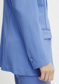 Niebieska marynarka o dopasowanym kroju z gładką fakturą, z detalem guzikowym na mankiecie oraz czystymi liniami wzdłuż rękawów i kołnierza.