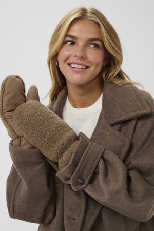 Femme souriante portant un manteau en laine brun et des moufles assorties en laine pelucheuse, regardant légèrement sur le côté sur un fond gris clair uni.