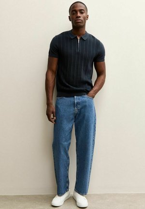 Mężczyzna stojący oparty o jasną ścianę, ubrany w czarną koszulkę z krótkim rękawem i zamkiem przy kołnierzu, niebieskie dżinsy i białe trampki, z jedną ręką w kieszeni.