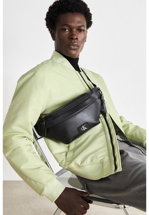 Jeune homme portant une veste vert clair et un sac bandoulière noir assis sur une chaise en métal devant un fond blanc.