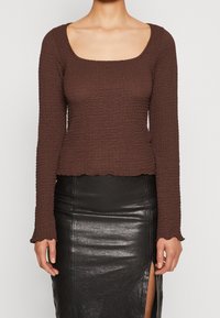 Brun långärmad topp med en strukturiserad, ribbad design och fyrkantig halsringning. Den har en kortad passform och vågiga fållar. Matchad med en svart kjol.