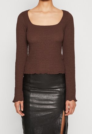 Braunes Langarmshirt mit strukturiertem, geripptem Design und quadratischem Ausschnitt. Es hat einen cropped Schnitt und gewellte Säume. Kombiniert mit einem schwarzen Rock.