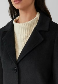 Schwarzer Wollmantel mit Reverskragen, Ein-Knopf-Verschluss und strukturiertem cremefarbenem Strickpullover darunter.