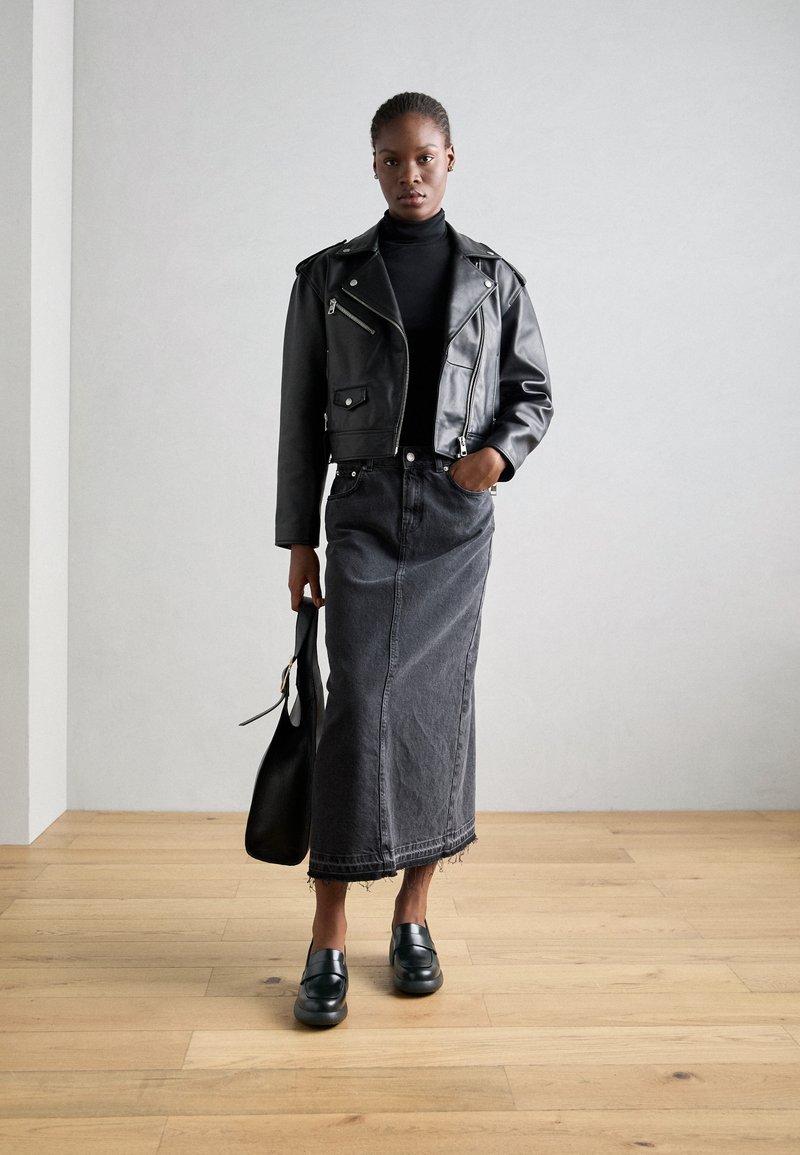 Veste de motard en cuir noir sur un col roulé noir, associée à une longue jupe en denim grise et des mocassins noirs, tenant un sac à main noir.