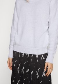 Maglione lavorato a maglia grigio chiaro con orlo a coste, abbinato a una gonna nera plissettata con motivi astratti bianchi e una vita aderente.